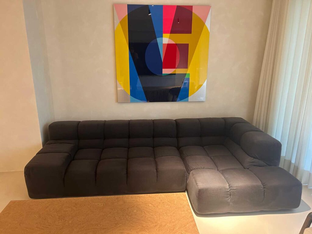 Sofá seccional oscuro en una sala moderna con una pintura abstracta de fondo. Realizar una limpieza profesional del sofá de forma regular ayuda a mantenerlo en perfecto estado, eliminando la suciedad acumulada y prolongando su vida útil, especialmente en hogares con uso frecuente.